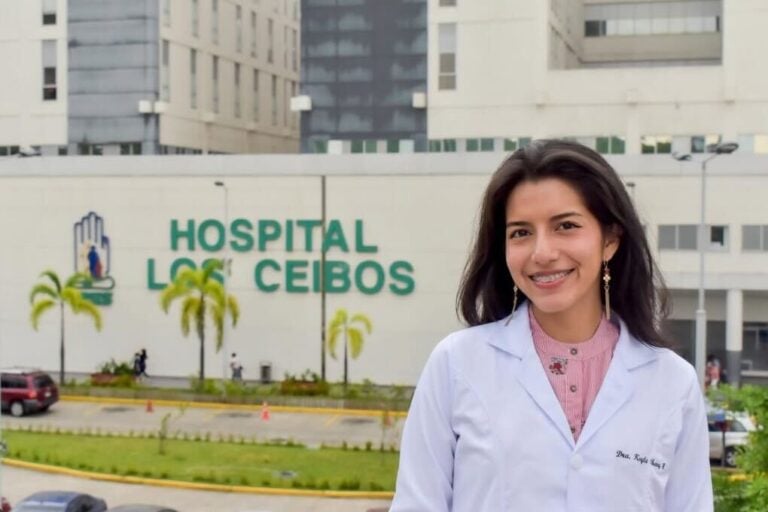 Medical professional standing outside a hospital in Ecuador, wearing a white lab coat and smiling toward the camera.